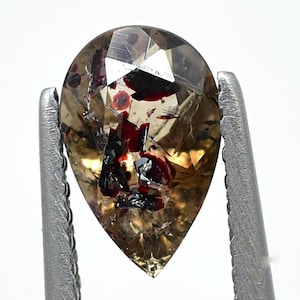 May include: A pear-shaped gemstone with a mix of brown, red, and clear facets, held by a silver tweezer. The gem has inclusions and a polished surface, showcasing a natural, earthy aesthetic. The gem is approximately 1 inch in length.