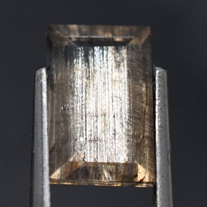 Puede incluir: Una gema de corte rectangular sujeta con pinzas. La gema es de color marrón claro con estrías verticales y forma rectangular con parte superior plana. Las pinzas son plateadas y el fondo es gris oscuro.
