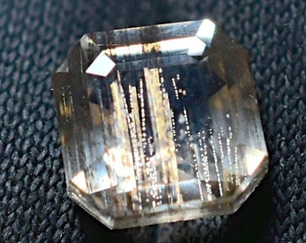 Puede incluir: Una gema de corte rectangular con un tono marrón claro y transparente. La gema tiene múltiples facetas que reflejan la luz, creando un efecto brillante. El interior presenta inclusiones lineales verticales. La gema está sobre un fondo oscuro y texturizado.