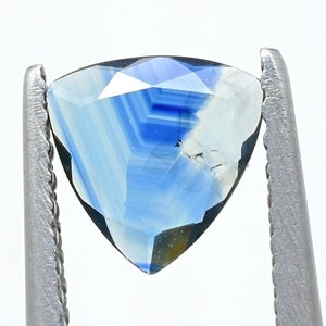 May include: A triangular gemstone with a gradient of blue and white hues. The gem is held by a silver metal tool, showcasing its faceted surfaces and internal patterns. The stone's shape is a perfect triangle, with a polished finish.