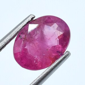 May include: A vibrant, oval-cut pink gemstone with a faceted surface that reflects light. The stone is held by a pair of silver-toned metal tweezers, showcasing its translucent quality and deep magenta hue against a plain, light-colored background.