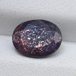 May include: An oval-cut gemstone with a dark purple base and shimmering inclusions. The facets reflect light, creating a sparkling effect. The gemstone is set against a white background. The stone's dimensions are in centimetres.