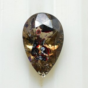 May include: A faceted, pear-shaped gemstone with a smoky brown hue. The gem features internal inclusions of dark red and black, creating a unique pattern. The stone is cut with multiple facets, enhancing its brilliance.