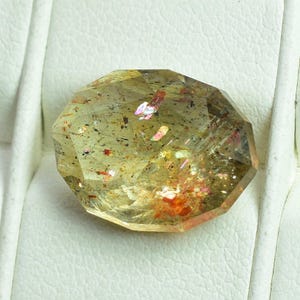 May include: A faceted gemstone with a light yellow hue, showcasing internal inclusions of red and iridescent colors. The gem is set against a white background, highlighting its clarity and unique characteristics. The gemstone is a unique piece.