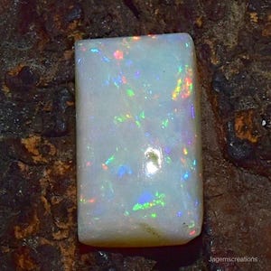 Könnte beinhalten: Ein rechteckiger, polierter Opal-Edelstein mit milchig-weißer Basis. Der Opal zeigt lebhafte Farbblitze, darunter Blau, Grün und Gelb, über seine Oberfläche. Der Edelstein ist vor einem dunklen, strukturierten Hintergrund platziert.