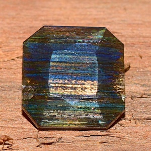 Könnte beinhalten: Ein facettierter Edelstein mit quadratischer Form und abgeschrägten Kanten. Der Stein zeigt eine Mischung aus blauen, grünen und goldenen Farben mit einem gestreiften Muster. Der Edelstein liegt auf einer Holzoberfläche.