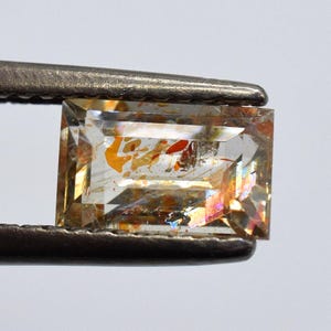 Puede incluir: Una gema de corte rectangular sujeta con pinzas. La gema tiene una base transparente con inclusiones internas de colores naranja, rojo e iridiscente. Las facetas reflejan la luz, creando un efecto brillante. Las pinzas son plateadas.