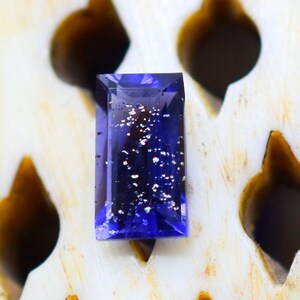 May include: A rectangular, faceted gemstone in a deep, translucent violet-blue hue. The stone features internal inclusions that appear as scattered, reflective silver-toned metallic flecks, creating a sparkling effect within the dark blue body.