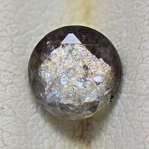 May include: A faceted, round gemstone with a dark, smoky gray hue. The gem has a reflective, iridescent surface with patches of gold and silver. The top facet is a small, triangular shape. The gemstone is set against a neutral background.
