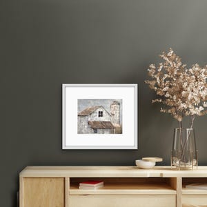 May include: A framed print of a weathered white farmhouse with a silo, set against a cloudy sky. The farmhouse has a single window and a brown roof.