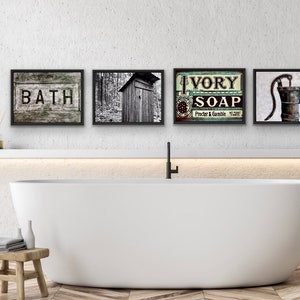 May include: Four framed vintage-style prints above a white bathtub. The prints feature the word "BATH", a black and white photo of an outhouse, an "IVORY SOAP" advertisement, and a water pump. The bathroom has a wooden stool and a green plant.
