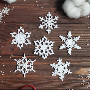 May include: Six white crocheted snowflakes on a brown wooden background. The snowflakes are arranged in a semi-circle with the points of the snowflakes facing inwards.