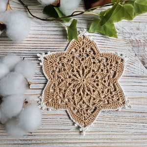 May include: A beige crocheted star-shaped doily with a white border. The doily is lying on a white wooden surface.