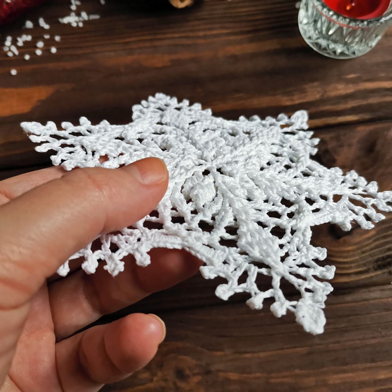 May include: A white crocheted snowflake with a delicate, intricate design. The snowflake is held in a hand against a wooden background.