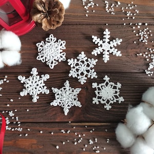 May include: Six white crocheted snowflakes of varying designs are arranged on a dark wooden surface. The snowflakes are intricately crafted, showcasing delicate patterns. Decorative elements like cotton bolls and a pine cone are also present, suggesting a winter or holiday theme.