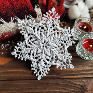 May include: A white crocheted snowflake ornament with intricate details. The snowflake is on a brown wooden surface with a red knitted blanket and candles in the background.