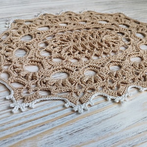May include: A beige crocheted doily with a white crocheted border. The doily has a circular design with a central motif and a repeating pattern of circles and loops.