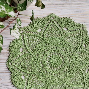 Peut inclure: Napperon au crochet fait à la main, vert clair, avec un motif ajouré détaillé. Le napperon circulaire présente un motif central en spirale et un bord festonné. La texture en dentelle complexe est visible, mettant en valeur le savoir-faire. Sur une surface en bois clair.