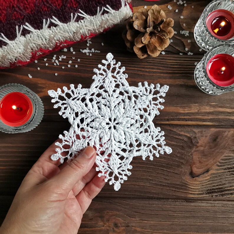 May include: White crocheted snowflake ornament on a wooden background. The snowflake is delicate and intricate, with a six-pointed star shape.