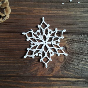 May include: A white, crocheted snowflake ornament with a six-pointed star design. The snowflake is displayed on a dark brown wooden surface. The ornament is a winter holiday decoration.