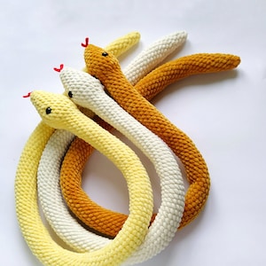 May include: Three crocheted snake toys in yellow, white and orange colours. The snakes are curled together in a circle.