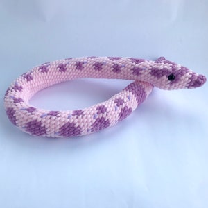 Peut inclure: Un jouet serpent en crochet rose et violet. Le serpent est enroulé en cercle avec sa tête reposant sur le sol.