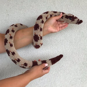 May include: A crocheted snake toy with brown and beige spots. The snake is curled up in a person's hands.