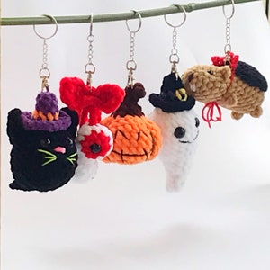 May include: Five crocheted Halloween-themed keychains hanging from silver chains. The keychains include a black cat wearing a purple hat, a red and white eyeball, an orange pumpkin, a white ghost wearing a black hat, and a brown and black hamster wearing a red and black hat.