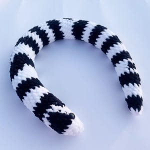 May include: A crocheted snake toy in a black and white pattern. The snake is curved, with a rounded head and a small, black bead for an eye. The body features a repeating chevron pattern in black and white.