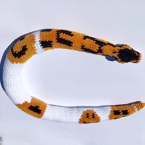 May include: A crocheted snake toy with a white base and brown and black spots. The snake is curled into a circle.