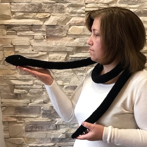 May include: A black crocheted scarf in the shape of a snake. The scarf is long and thin, and the person is holding it up to show its length.