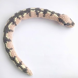 May include: A plush, crocheted snake toy in shades of gray, cream, and light pink. The snake is curved, with a detailed pattern mimicking scales. The head is slightly raised, and the eyes are visible. The toy is soft and appears handmade.