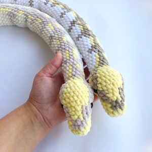 May include: Two crocheted snakes, one white and one yellow, with brown accents, are curled together. The snakes are made with a fuzzy yarn and have realistic details.
