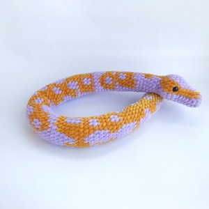 May include: A plush, crocheted snake toy in shades of orange and lavender. The snake's body is patterned with orange and white spots on a lavender background. The head features a small black eye. The toy is designed to be soft and cuddly.