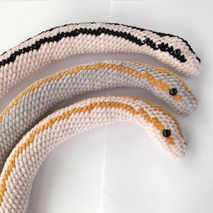May include: Three crocheted snake toys in different sizes. The largest snake is black and white, the middle snake is gray and gold, and the smallest snake is beige and gold.