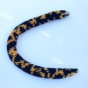 May include: A curved, crocheted item in black with a repeating pattern of yellow shapes. The item has a rounded shape and a slightly lighter, beige tip. The background is a plain, light blue surface.