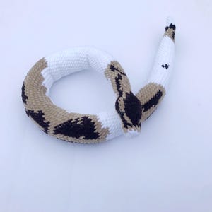 May include: A crocheted snake toy with brown, white, and black markings. The snake is curled in a circle with its head facing the camera.