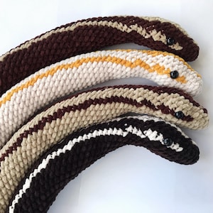 May include: Four handmade crochet snakes in various colours. The snakes have black button eyes and are in shades of brown, beige, and white. One snake has a yellow stripe, and another has a white stripe. The snakes are soft and plush.