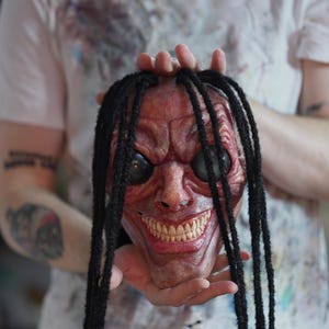 Puede incluir: Una máscara de rostro completo con un tono de piel rojizo rosado texturizado, arrugas faciales profundas, grandes ojos negros brillantes y una amplia sonrisa con dientes. La máscara está adornada con largos mechones de pelo negro texturizado.