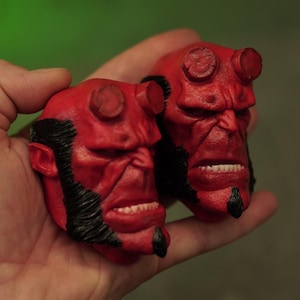 Puede incluir: Dos esculturas de cabezas rojas y detalladas de Hellboy con cabello y vello facial negro se sostienen en una mano. Cada cabeza tiene una expresión severa, cuernos y dientes visibles. El fondo es borroso, verde y gris.
