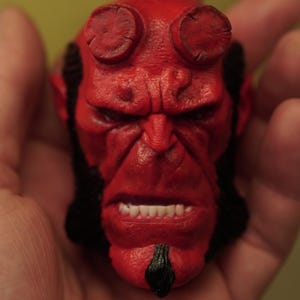 May include: A detailed, red figurine head with prominent horns and a stern expression. The figure has black hair and a goatee, with visible teeth. The overall design suggests a character from a comic book or fantasy genre.
