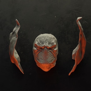 May include: A grey sculpted mask featuring a textured, furrowed brow and a central nose ridge, flanked by two separate, elongated, flowing grey sculpted pieces that curve outward, all displaying a subtle orange glow along the lower edges.