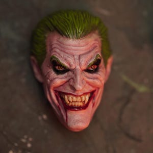 Könnte beinhalten: Eine detaillierte Skulptur des Gesichts des Jokers, mit grünen Haaren, einem blassen Teint und einem finsteren Grinsen. Die Augen sind dunkel mit roten Iris, und der Mund ist rot gefärbt. Das Design fängt das ikonische Aussehen der Figur ein.