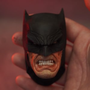 May include: A detailed, black Batman cowl with an open mouth revealing teeth. The face is painted with realistic skin tones, and the cowl has pointed ears. The image is set against a red background.