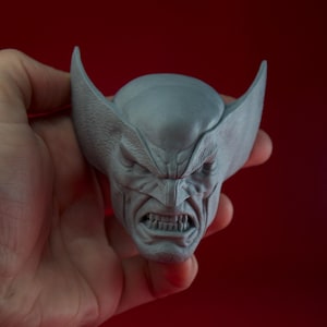 May include: A grey bust of a character with a fierce expression, featuring prominent brow ridges, sharp teeth, and large, pointed ears. The sculpture is held against a red background.