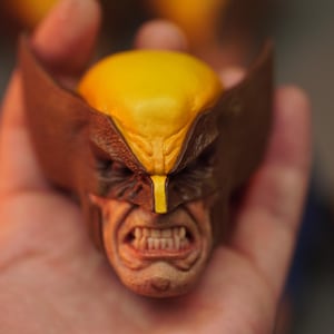 Peut inclure: Une figurine de tête de Wolverine miniature et détaillée. La figurine présente une cagoule jaune, un masque marron et une expression renfrognée avec des dents visibles. La figurine est tenue dans une main, mettant en valeur les détails complexes.