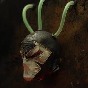 May include: A detailed close-up of a Bane mask, featuring a black and silver design with a distinctive faceplate. The mask has three green tubes attached, and is set against a textured, dark background.