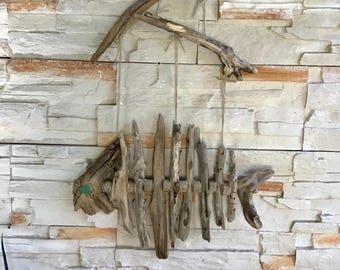 Può includere: Scultura rustica a forma di pesce in legno di deriva appesa a un muro di pietra. L'opera presenta un design a scheletro di pesce realizzato con pezzi di legno stagionato, con un piccolo accento verde. La scultura è sospesa con spago.