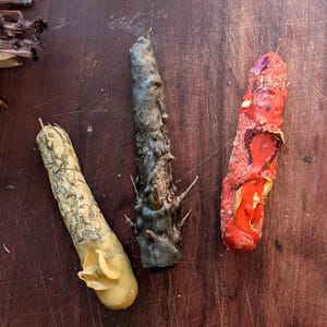 May include: Three decorative candles in a rustic setting. One candle is yellow and green, another is dark gray with spiky protrusions, and the third is red and orange. Dried flowers and a small silver container are also visible.