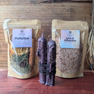 May include: Two stand-up pouches with clear windows and two purple taper candles tied together with black string. The left pouch is labelled Protection Simmer Pot Mix with dried orange, cinnamon, and herbs. The right pouch is labelled Love & Attraction Sweeping Salt.